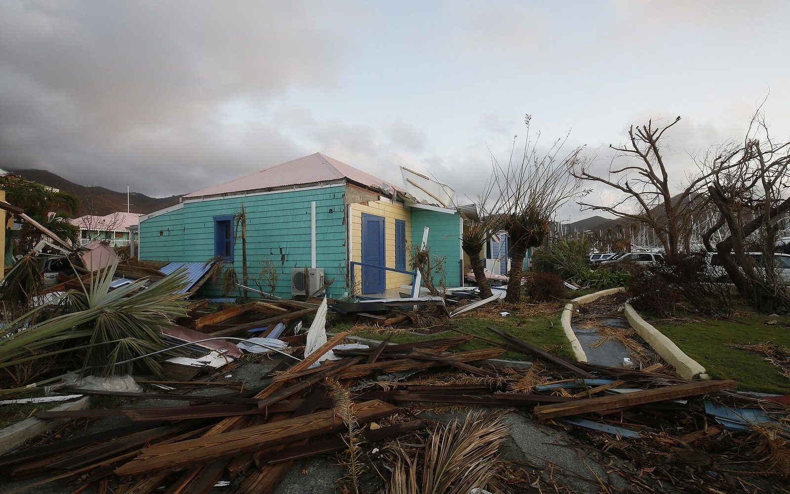 Wrecked debris and stripped trees surround a damaged turquoise and yellow house after a hurricane