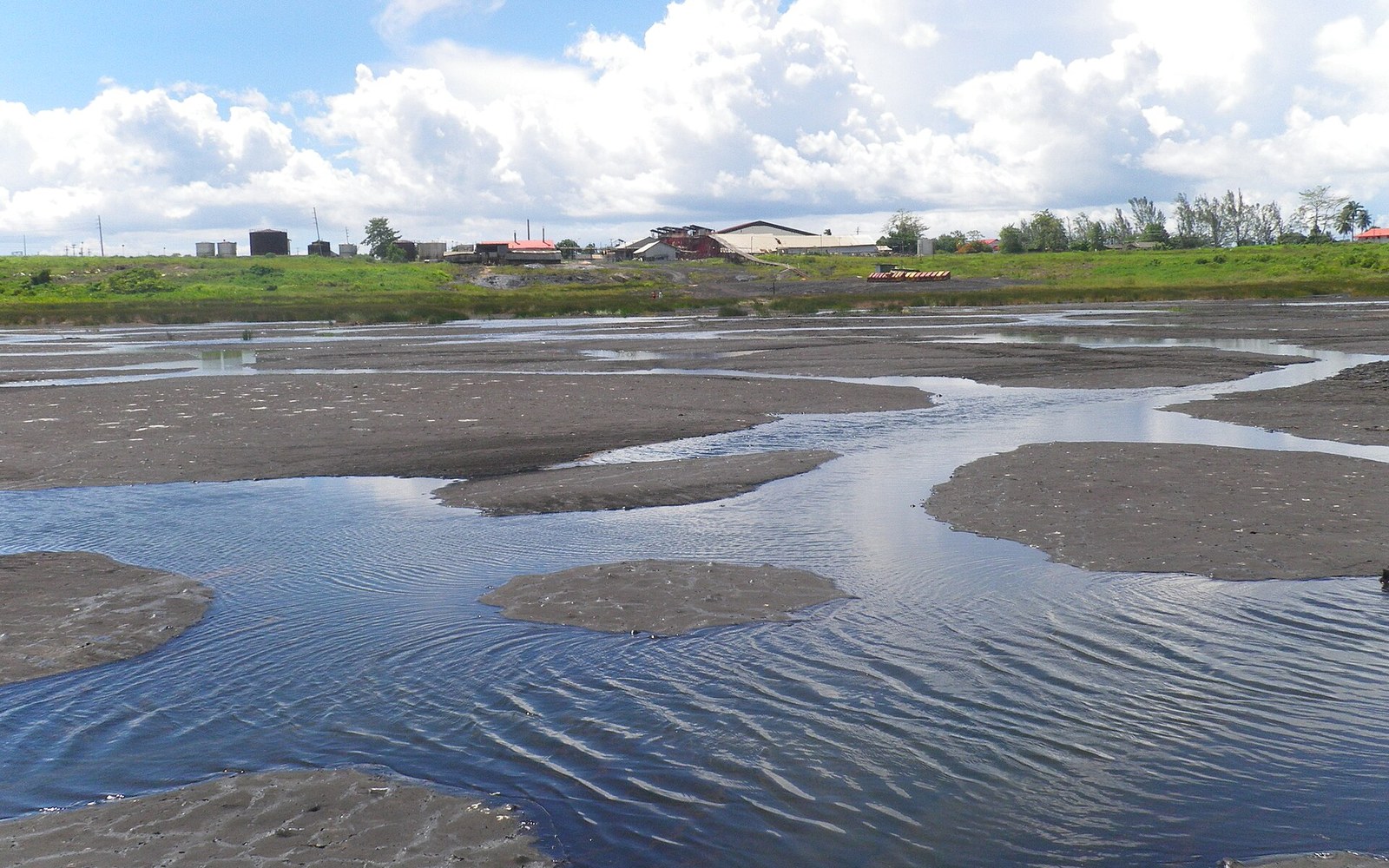 Shallow water pools and mud flats spread across Pitch Lake with buildings in the distance