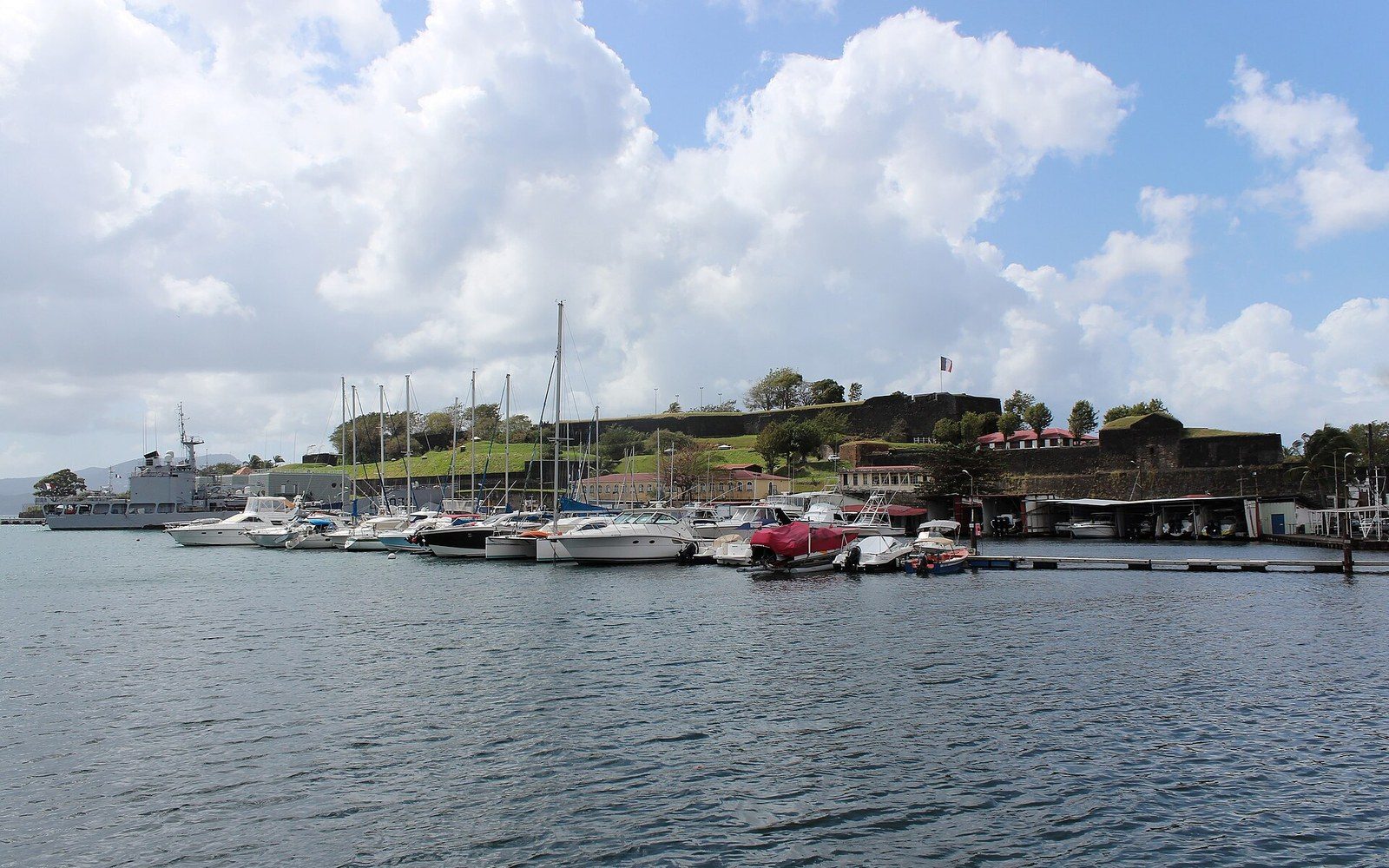 Yachts and a naval vessel moored near Fort Saint-Louis under a cloudy sky